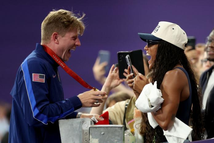 A male athlete shows his gold medal to a woman in the stands. She is holding her smartphone 